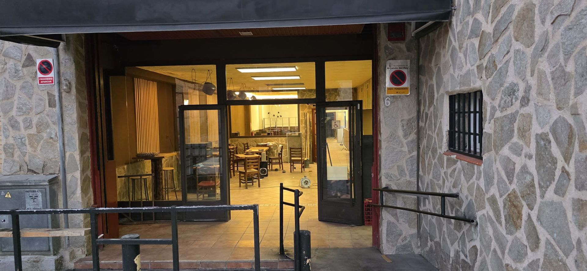 La entrada de la tienda tiene paredes de piedra, una puerta de cristal abierta y barandillas de metal que dan a una zona de estar interior.