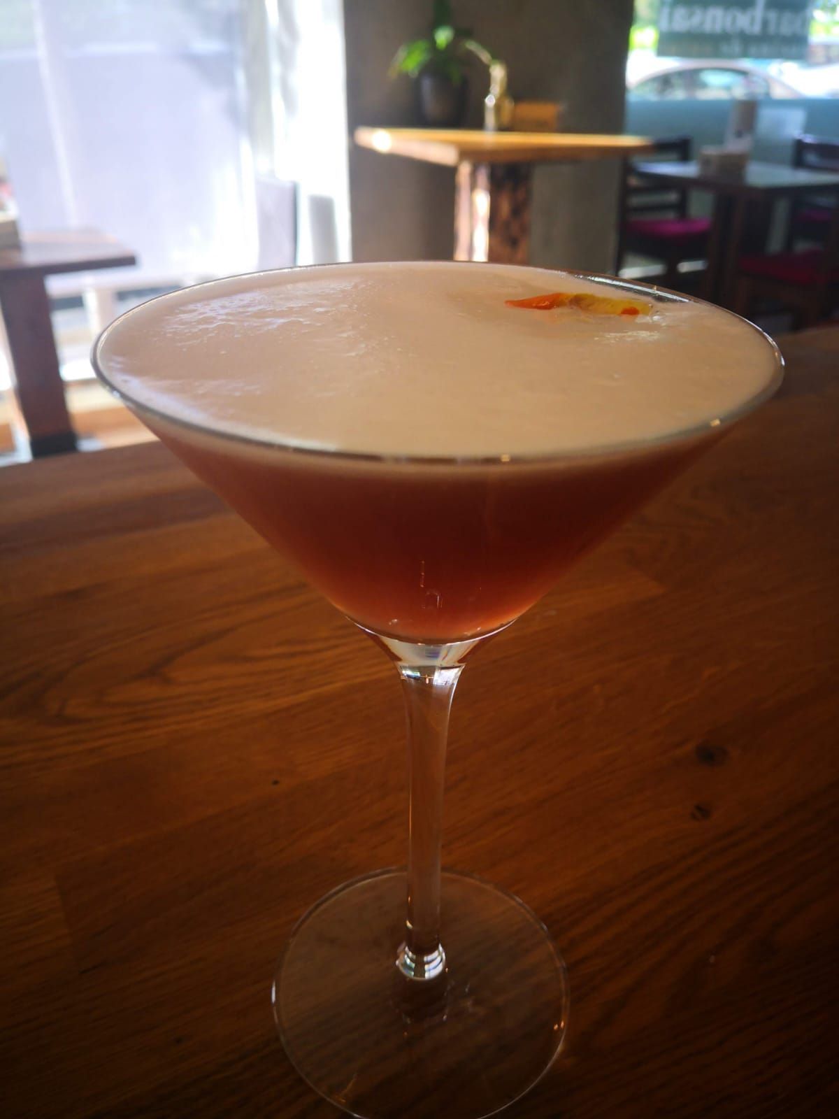 Un cóctel con una espesa y cremosa capa de espuma y una pequeña rodaja de naranja como guarnición, servido en una copa de martini sobre una mesa de madera.