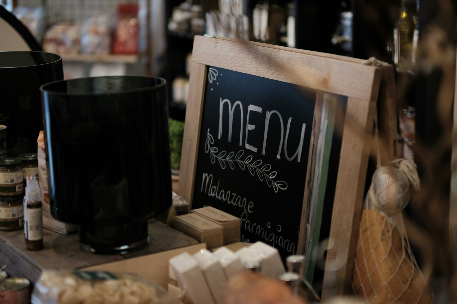 Un menú escrito en una pizarra con marco de madera, colocado sobre el mostrador de una tienda junto a un gran recipiente cilíndrico negro.