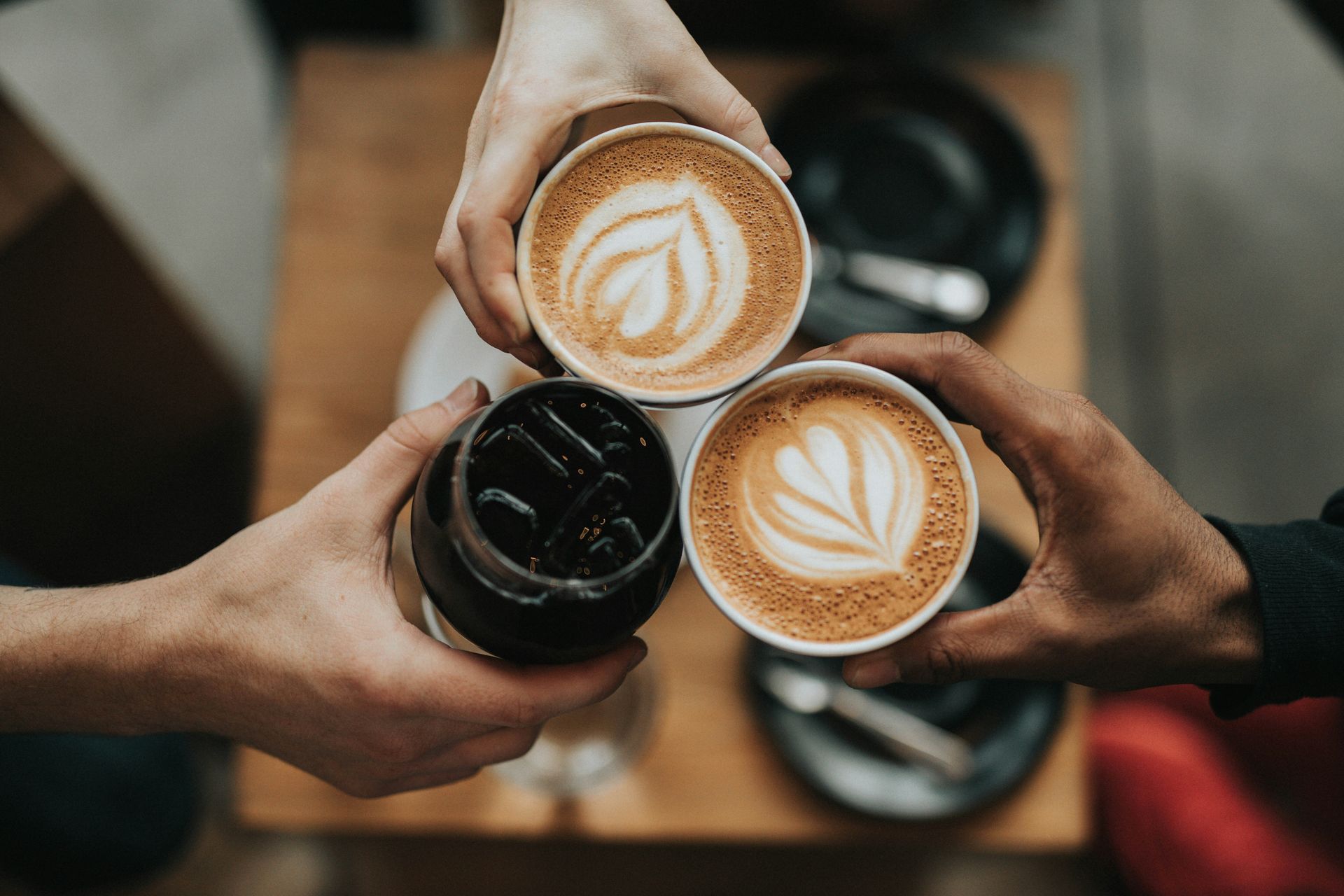 Tres personas brindando con dos cafés con arte latte y una bebida helada sobre una mesa de madera.