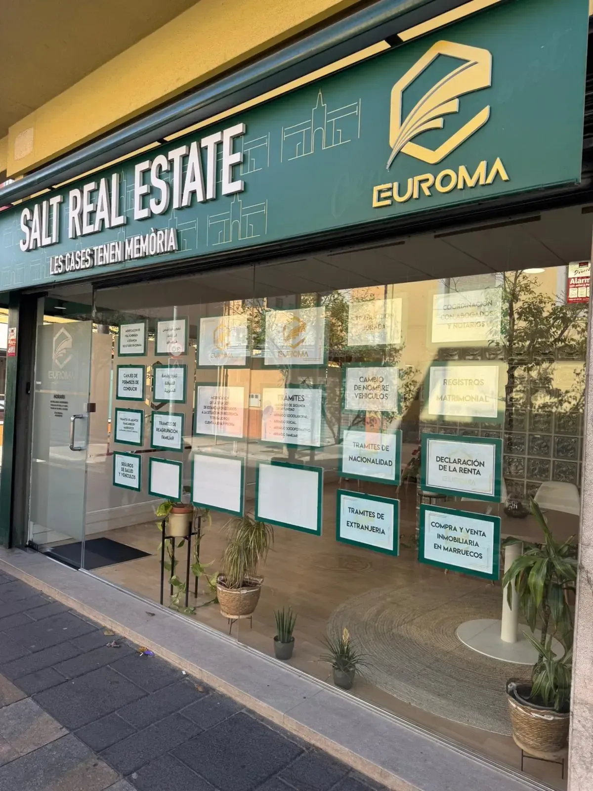 Local comercial de Salt Real Estate con anuncios. Letreros verdes y blancos. Escaparate con plantas en macetas.