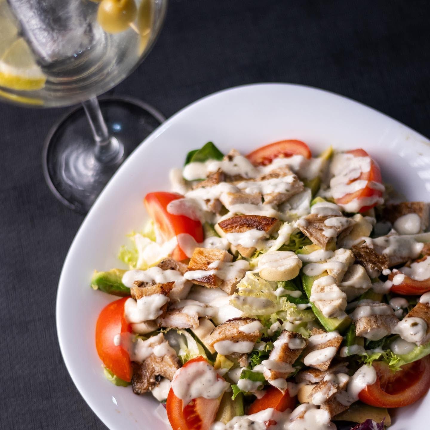 Ensalada con tomates, aguacate y pollo a la parrilla aderezada con vinagreta, acompañada de una copa de vino.