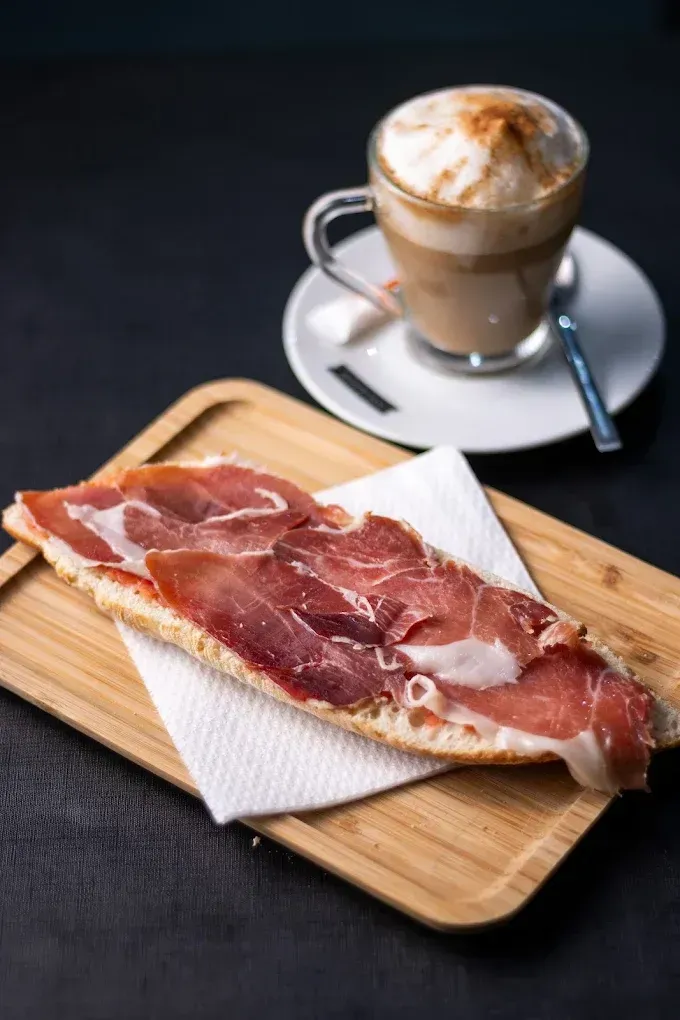 Una loncha de jamón curado sobre una tabla de madera junto a un capuchino en un platillo con cuchara.