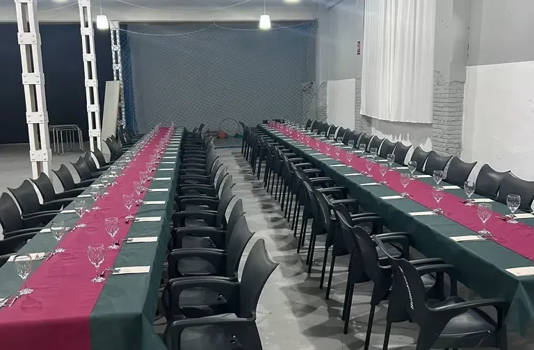 Filas de sillas negras junto a largas mesas de banquete con caminos de mesa rojos en un salón de eventos interior.