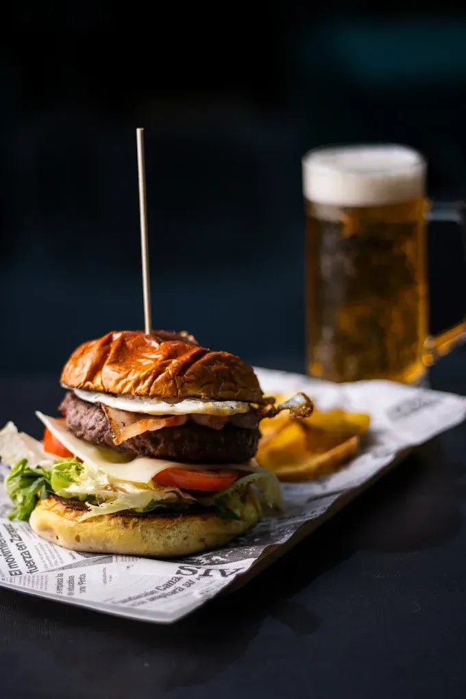 Hamburguesa gourmet con patatas fritas y una cerveza en una bandeja forrada de papel, iluminada contra un fondo oscuro.