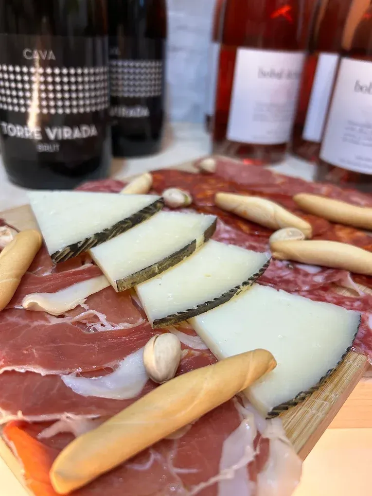 Aperitivo de queso y jamón serrano con palitos de pan sobre una tabla de madera, botellas de vino difuminadas al fondo.