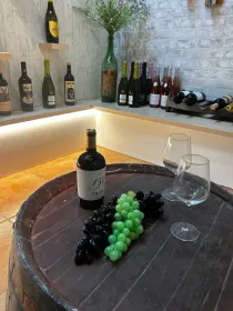 Exposición de vinos con uvas y una botella sobre una mesa rústica en una bodega con iluminación tenue.