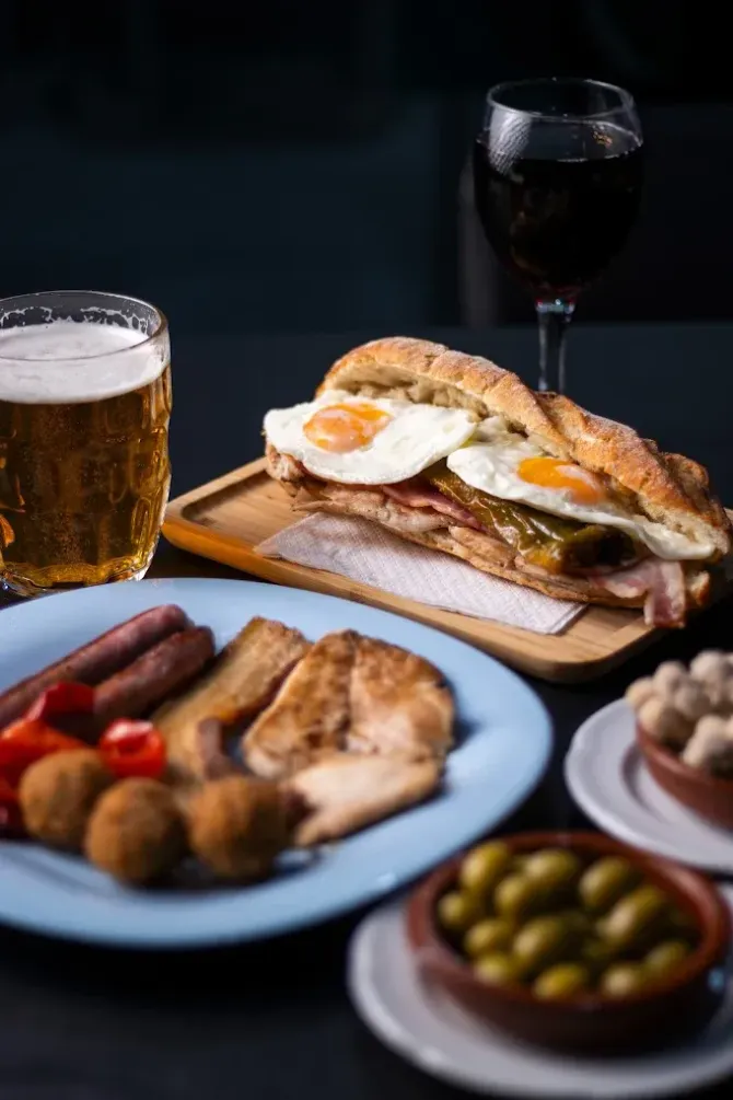 Mesa con cerveza, vino, sándwich, aceitunas y tapas sobre fondo oscuro.