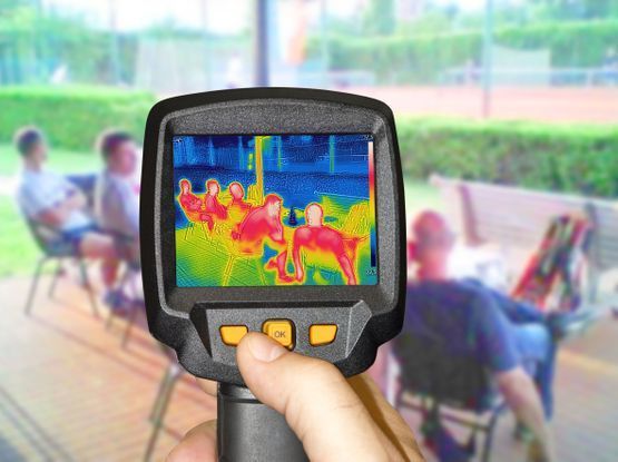 Una persona sostiene una cámara térmica frente a un grupo de personas sentadas en un banco.