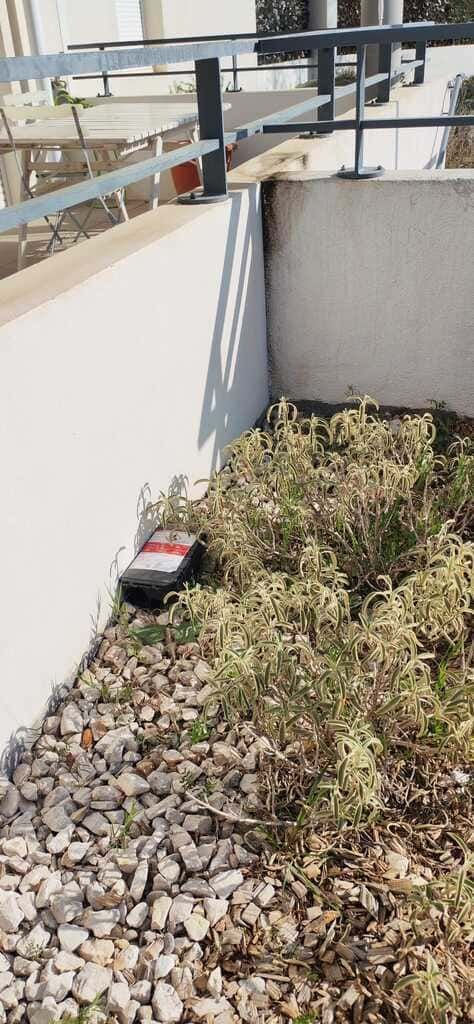 Coin de toit-terrasse avec du gravier, des mauvaises herbes, un mur blanc et un petit objet rouge et noir au sol