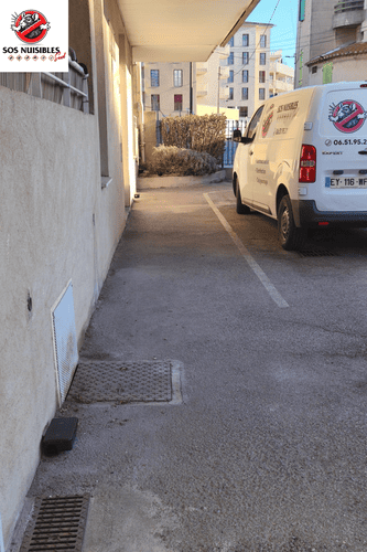 Une ruelle étroite longe un bâtiment, avec une camionnette blanche garée sur le côté droit.