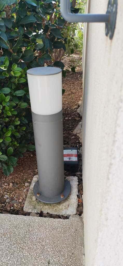 Poteau utilitaire gris près du mur d’un bâtiment, scellé dans une base en béton au milieu d’un aménagement paysager en gravier.