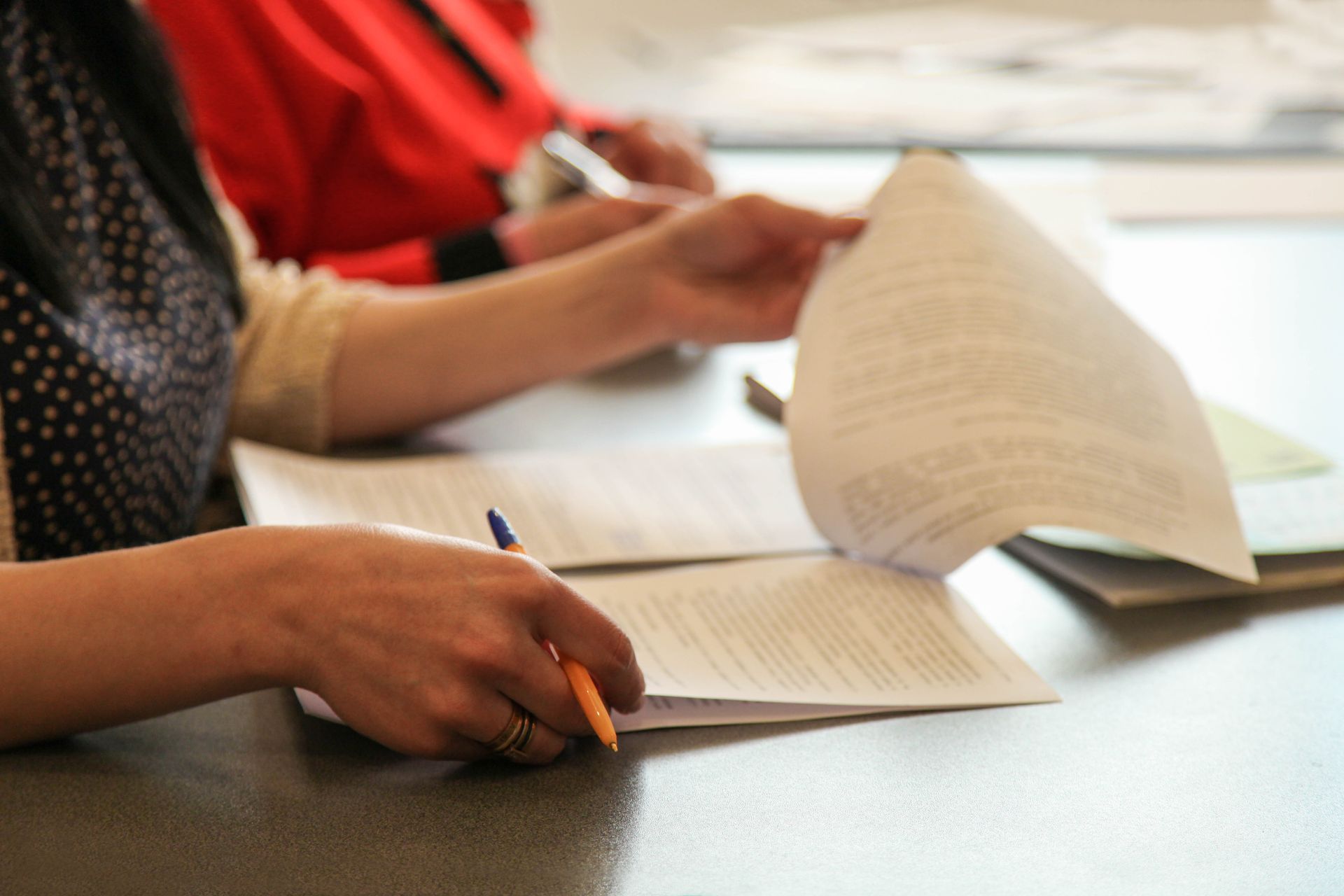 Une personne remet des documents à corriger sur une table, une page est tournée et un crayon se trouve à proximité.