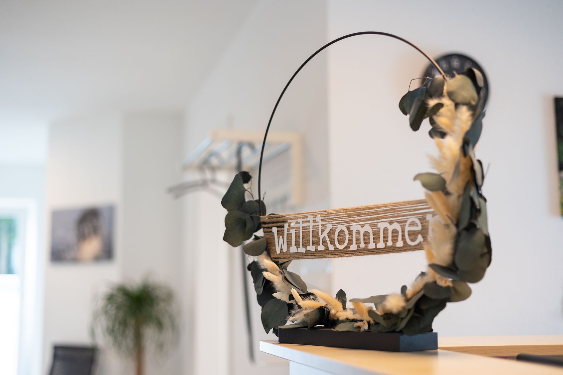 Willkommensschild in deutscher Sprache mit dekorativen Blättern auf einer weißen Theke. Helle Inneneinrichtung.