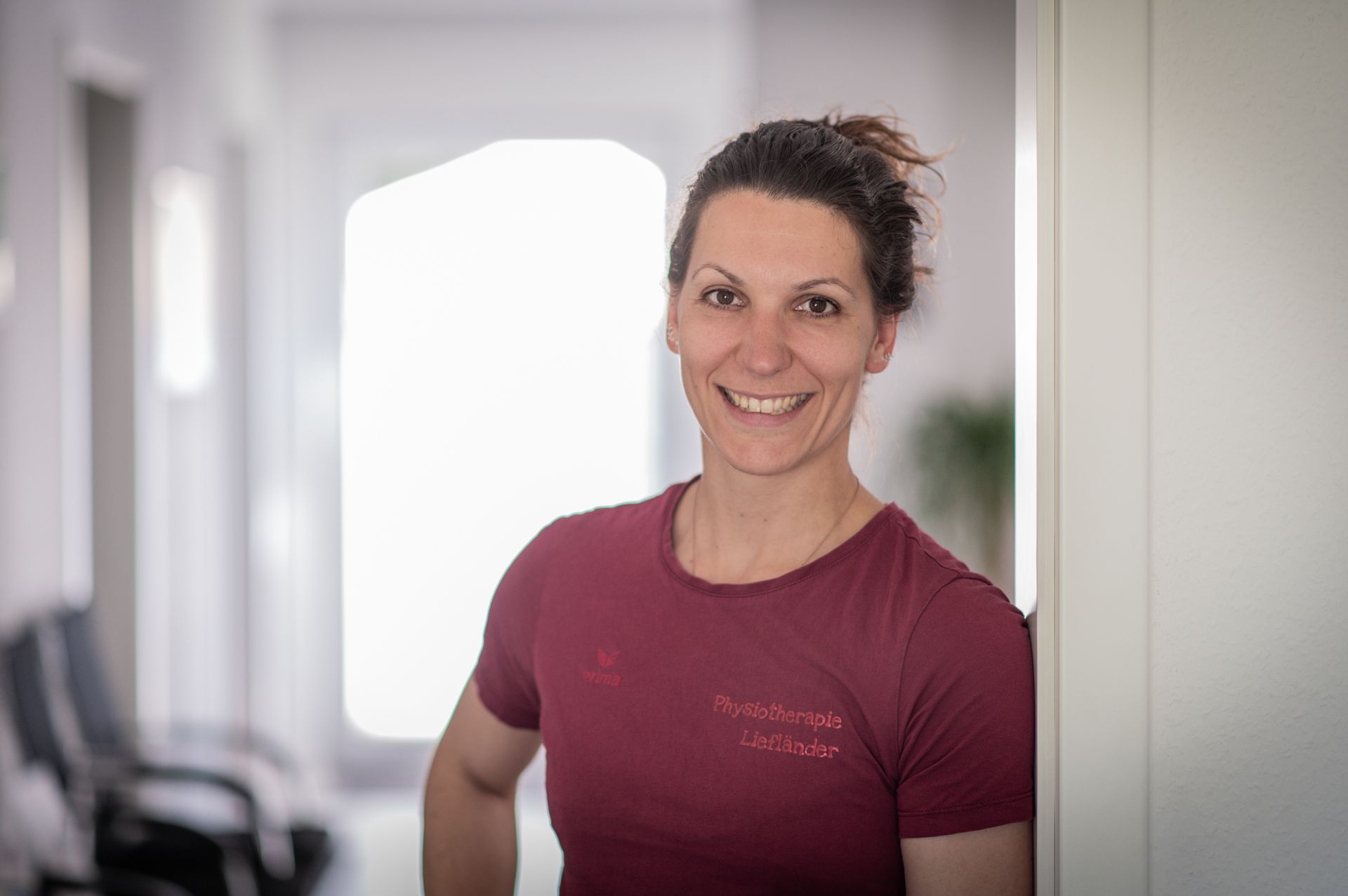 Eine Frau in einem burgunderfarbenen T-Shirt lehnt lächelnd in einem Türrahmen. Sie hat braunes Haar und befindet sich in einem Büro, in dem Stühle zu sehen sind.