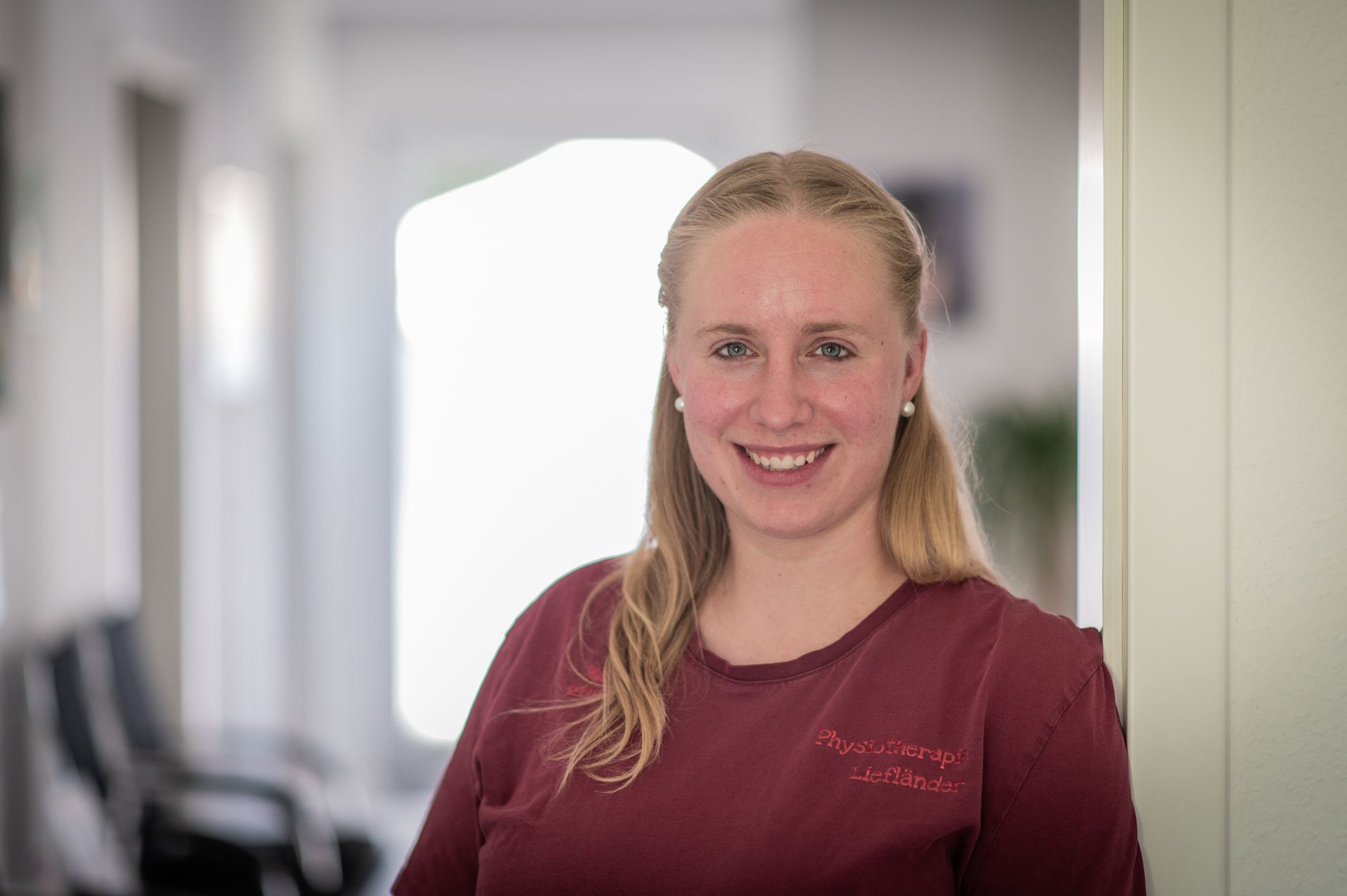 Blonde Frau in kastanienbraunem Hemd lächelt in einem Flur in die Kamera.