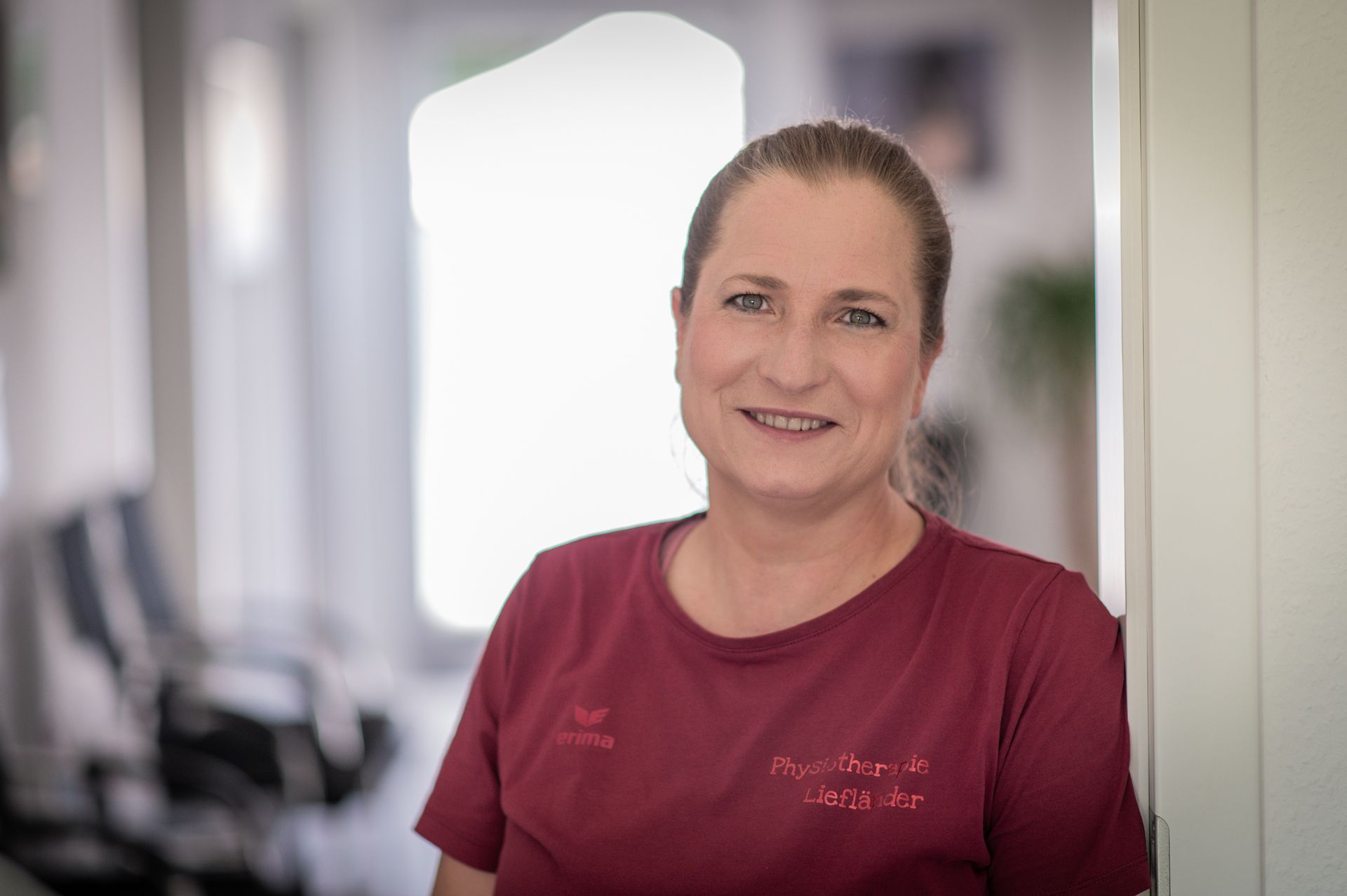 Lächelnde Frau in rotem T-Shirt, die in einem Büroflur an einem weißen Türrahmen lehnt.