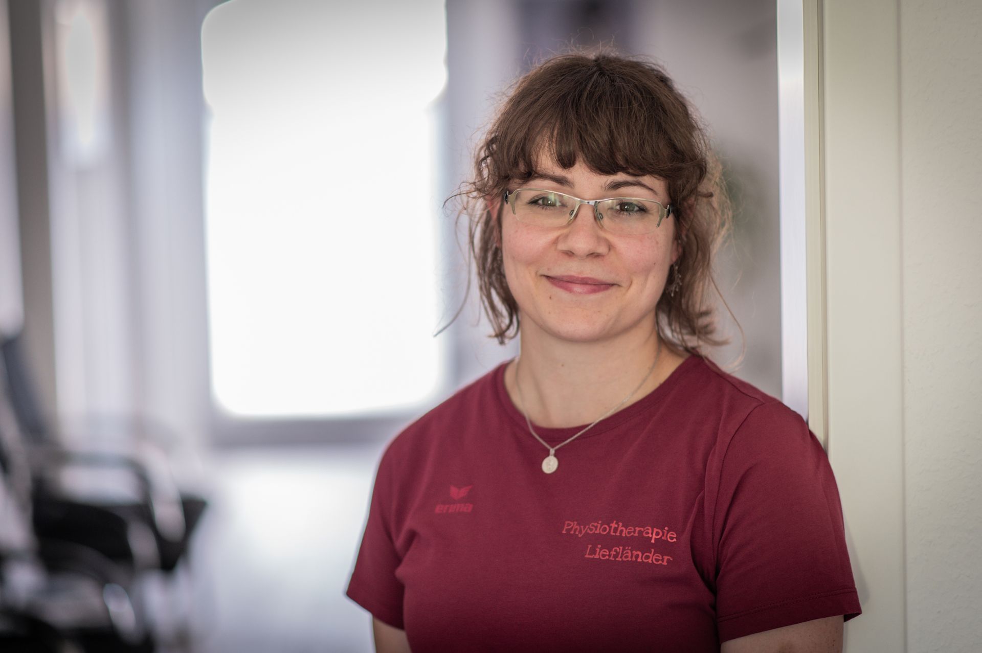 Eine lächelnde Frau mit Brille und kastanienbraunem Hemd steht in einem Flur. Natürliches Licht erhellt die Szene.