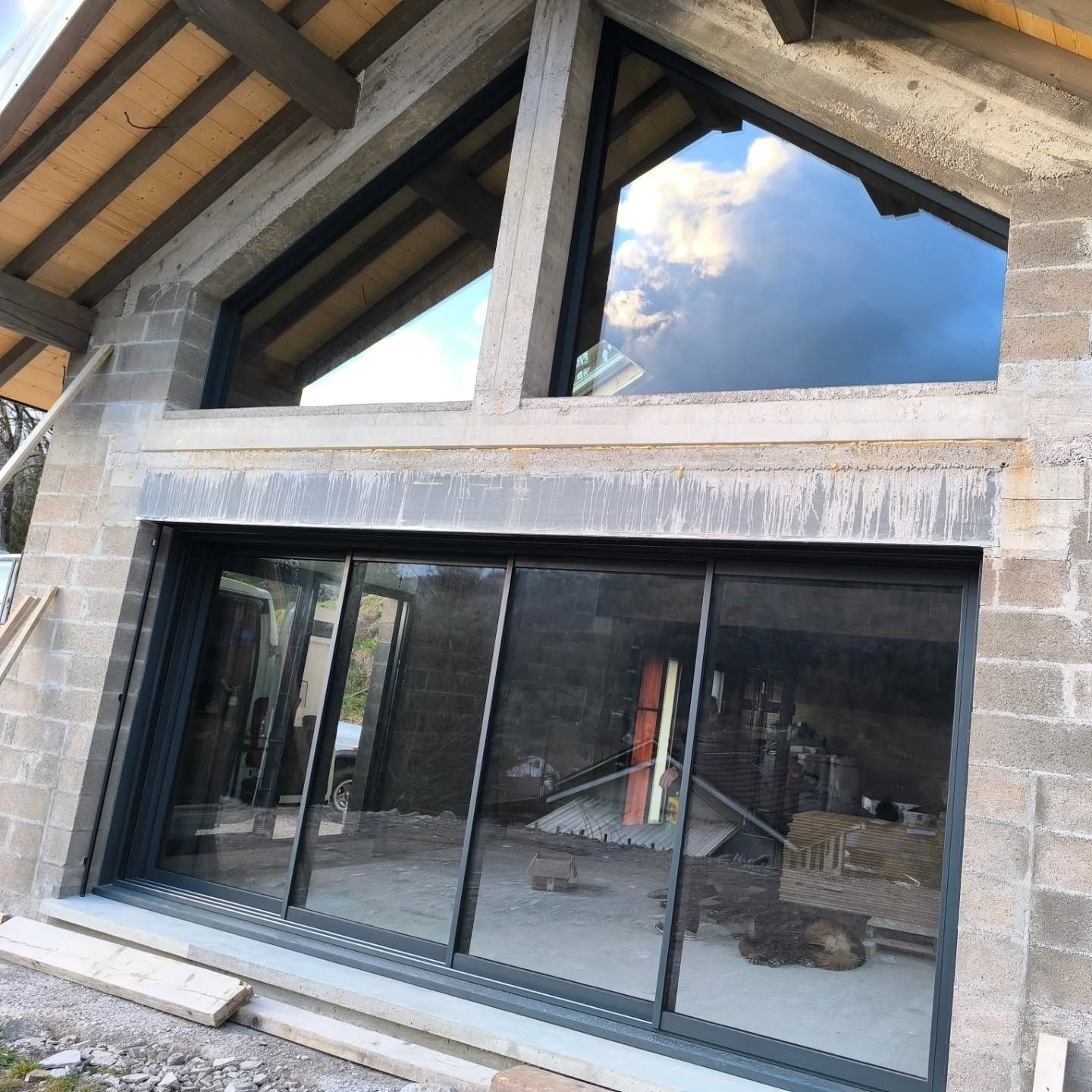 Un chantier de construction montrant une grande paroi vitrée à cadre sombre et deux fenêtres triangulaires intégrées à un mur en parpaings.