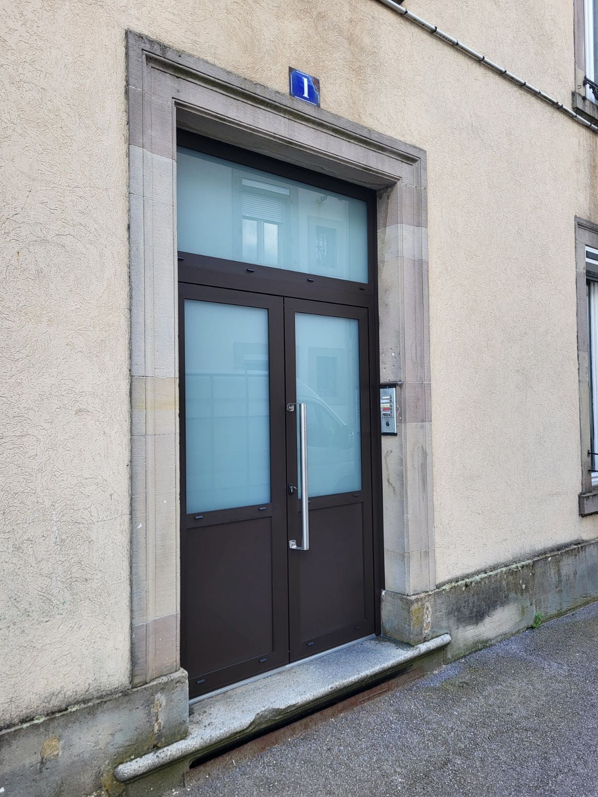 L'entrée d'un immeuble avec une double porte marron en aluminium, des panneaux de verre dépoli et un cadre en pierre surmonté du numéro 1.
