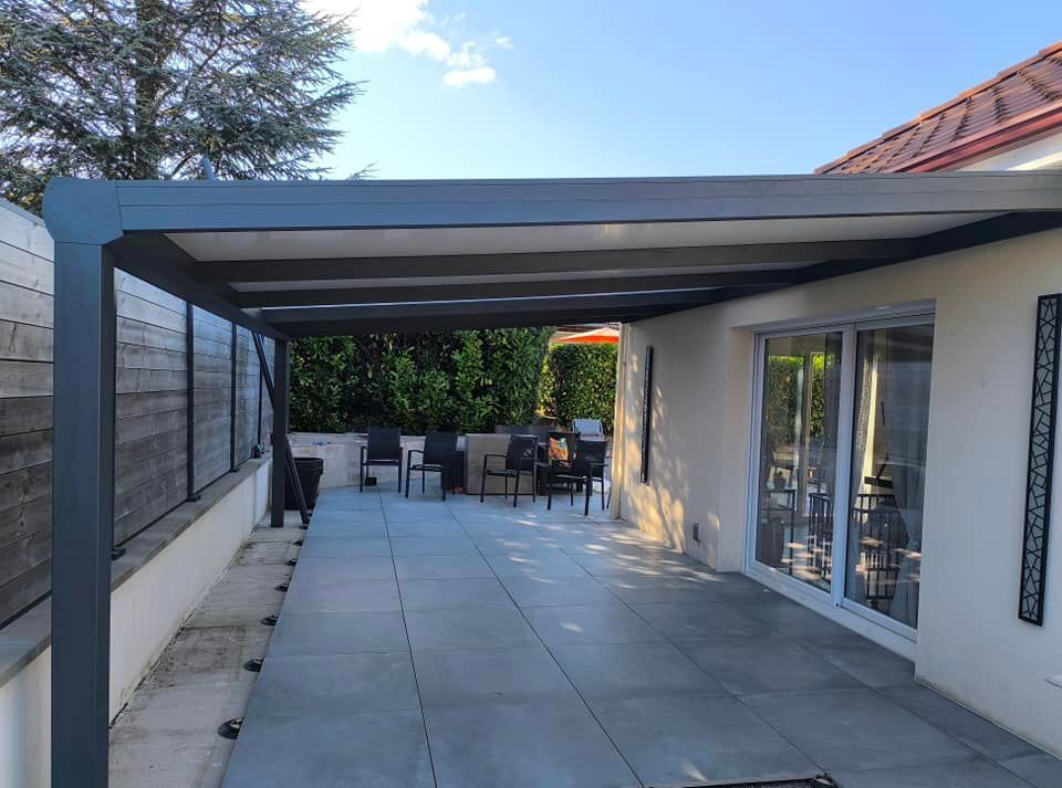 Une pergola en aluminium de couleur grise prolongeant une maison au-dessus d'une terrasse carrelée, avec du mobilier de jardin en arrière-plan.