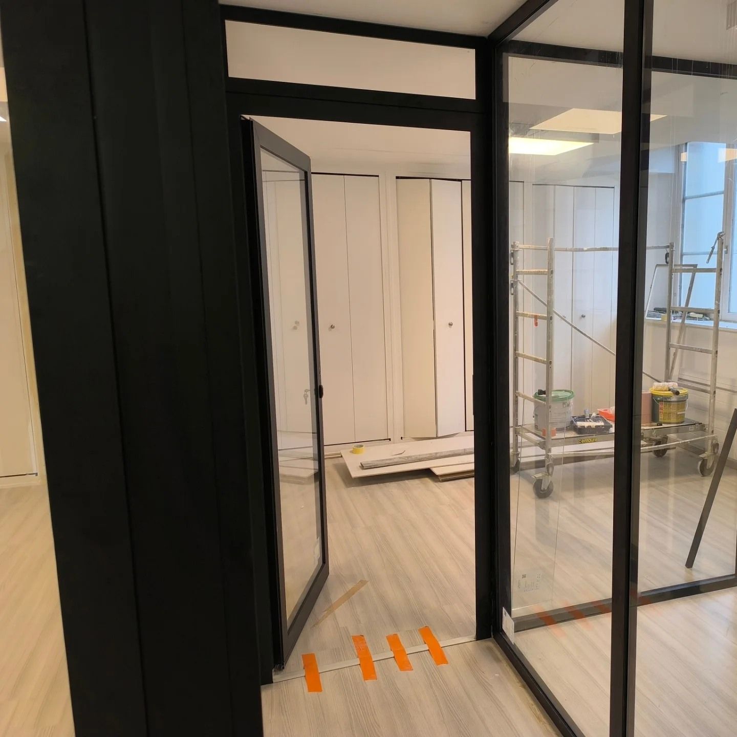 Un bureau en rénovation avec une porte vitrée, un parquet clair, des marquages au sol orange et des échafaudages à l'arrière.