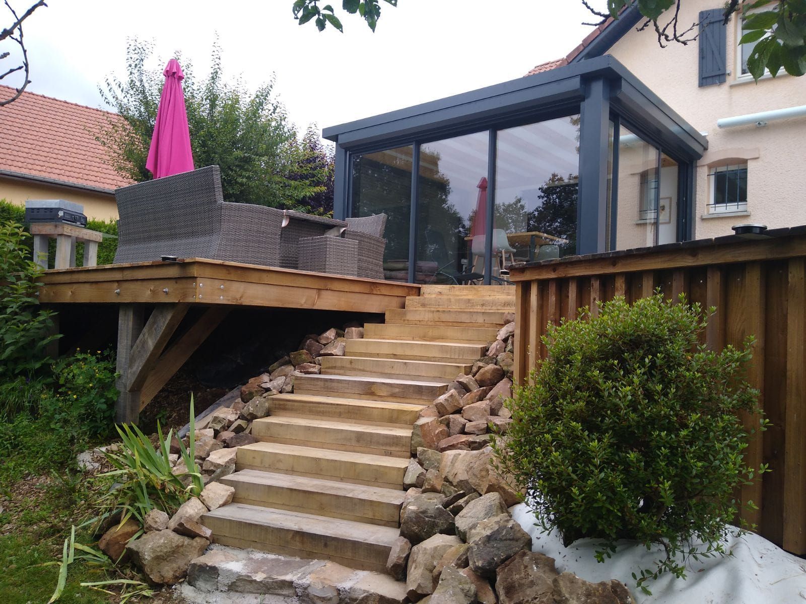 Un escalier en pierre mène à une terrasse en bois et à une véranda aluminium vitré attenant à la maison, avec des sièges extérieurs à proximité.