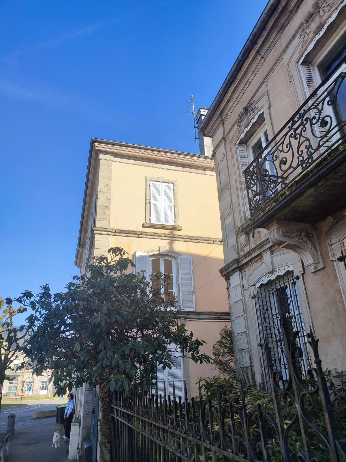 Vue ensoleillée de deux bâtiments historiques en pierre, avec volets alu blanc et balcons en fer forgé, sous un ciel bleu limpide.