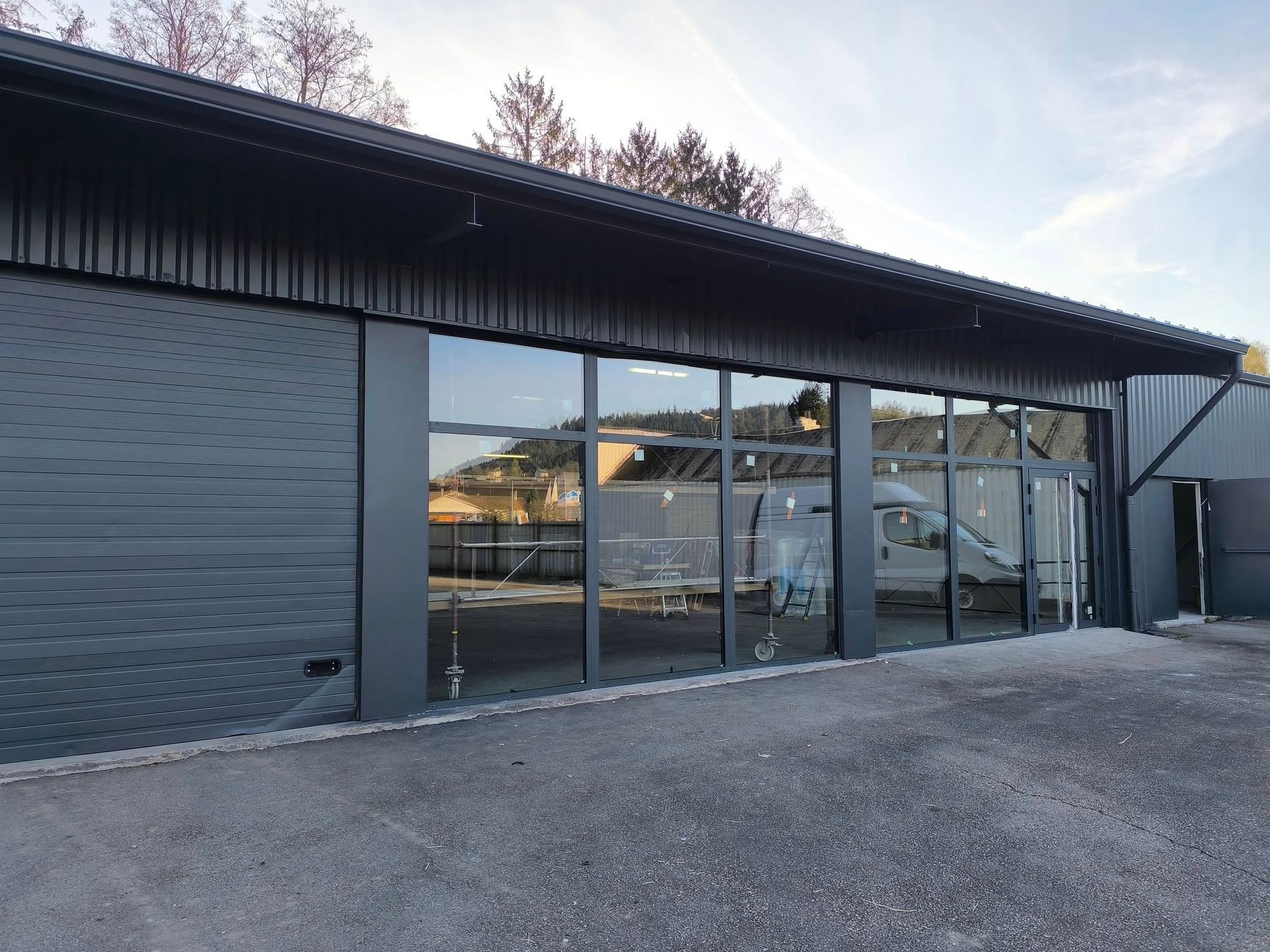 L'extérieur d'un bâtiment commercial se caractérise par de grandes baies vitrées, une façade en tôle ondulée sombre et un sol gravillonné.