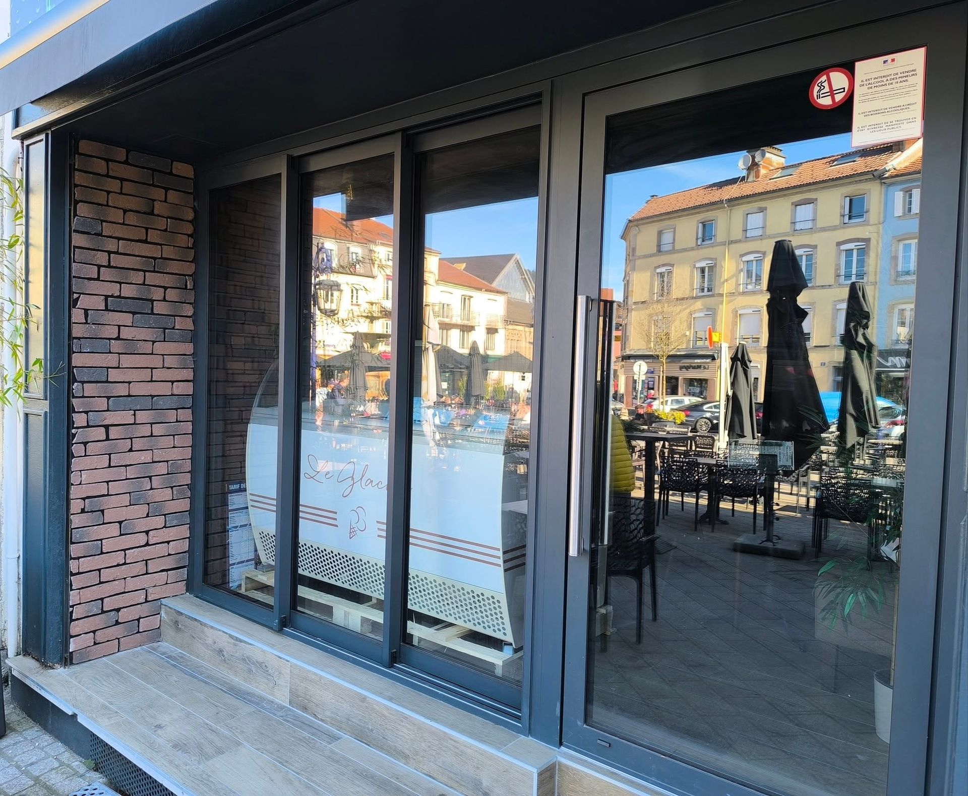 Façade commerciale avec revêtement en briques et portes vitrées, reflétant une scène de rue et une terrasse de restaurant.