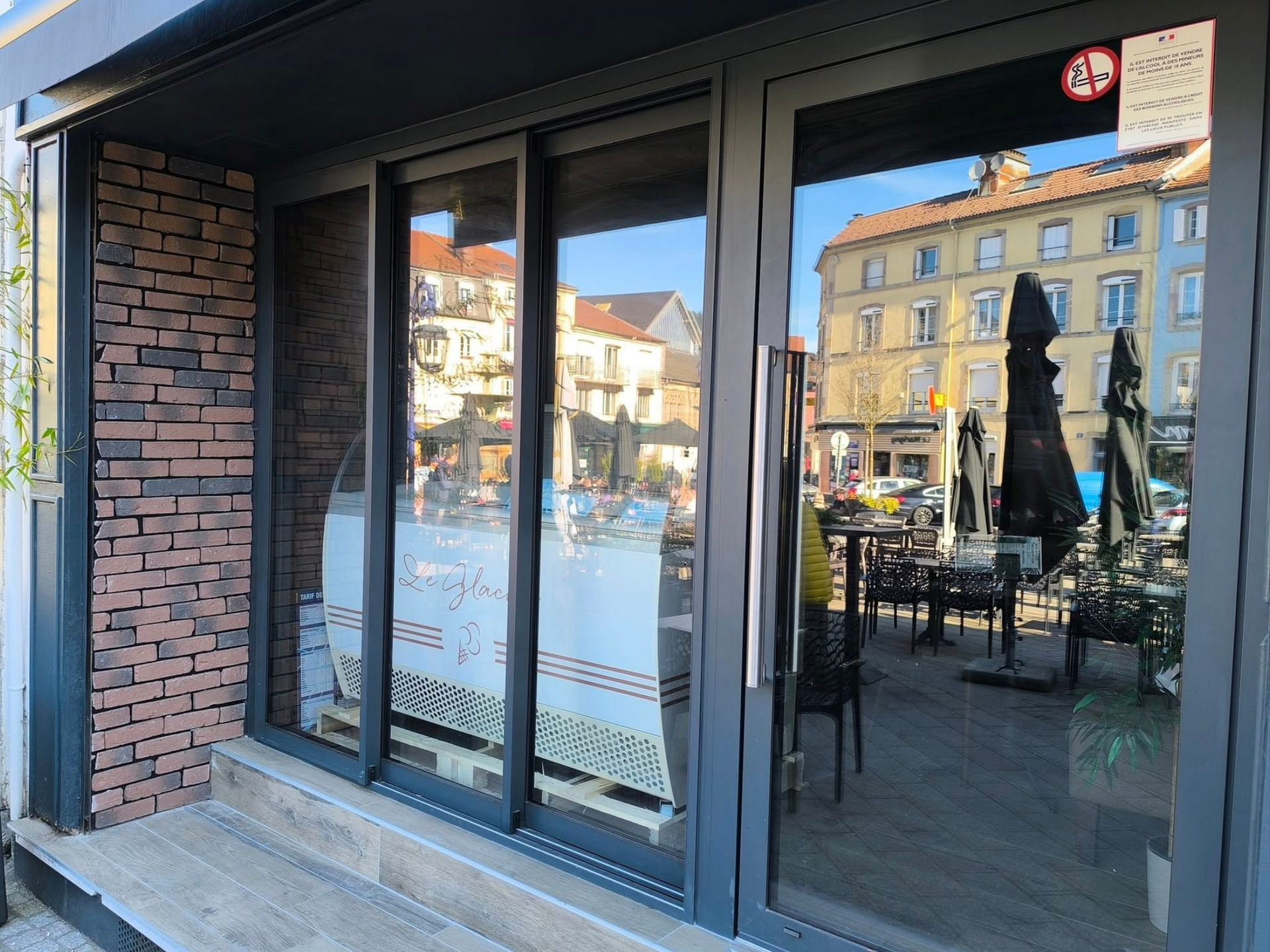 Façade de magasin avec un mur latéral en briques, de grandes portes coulissantes en aluminium et une vue réfléchissante sur une terrasse de café.
