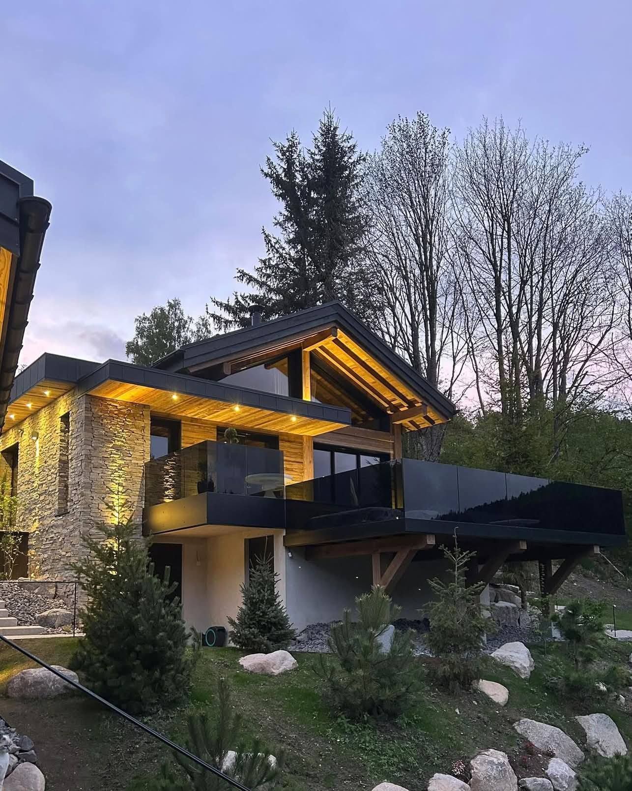 Une maison moderne aux murs de pierre et dotée d'un grand balcon sombre, éclairé par une lumière chaude au crépuscule, près d'une colline boisée.