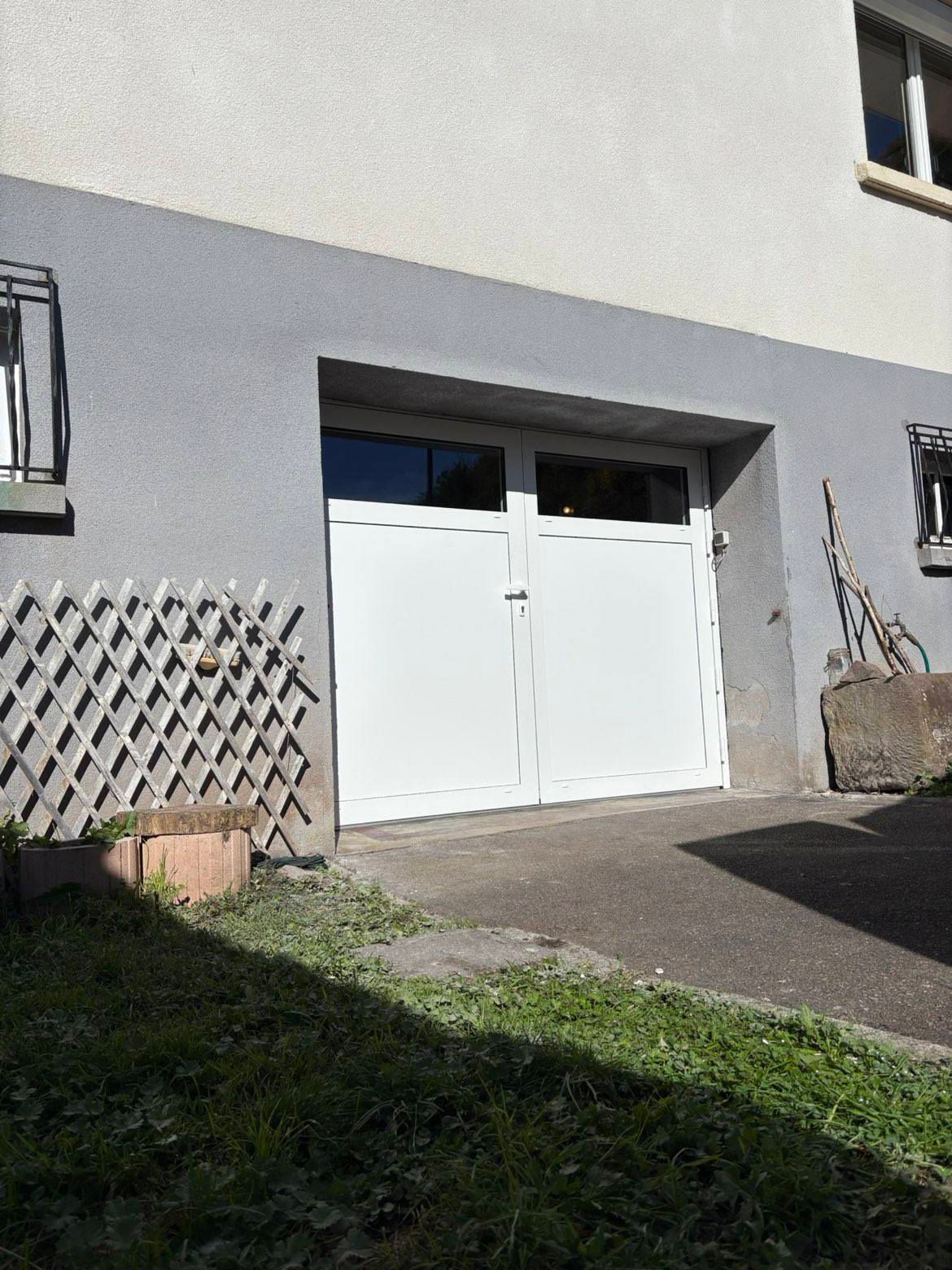 Un garage blanc à double porte, encastré dans un mur extérieur, avec un treillis en bois sur la gauche et un sol pavé.