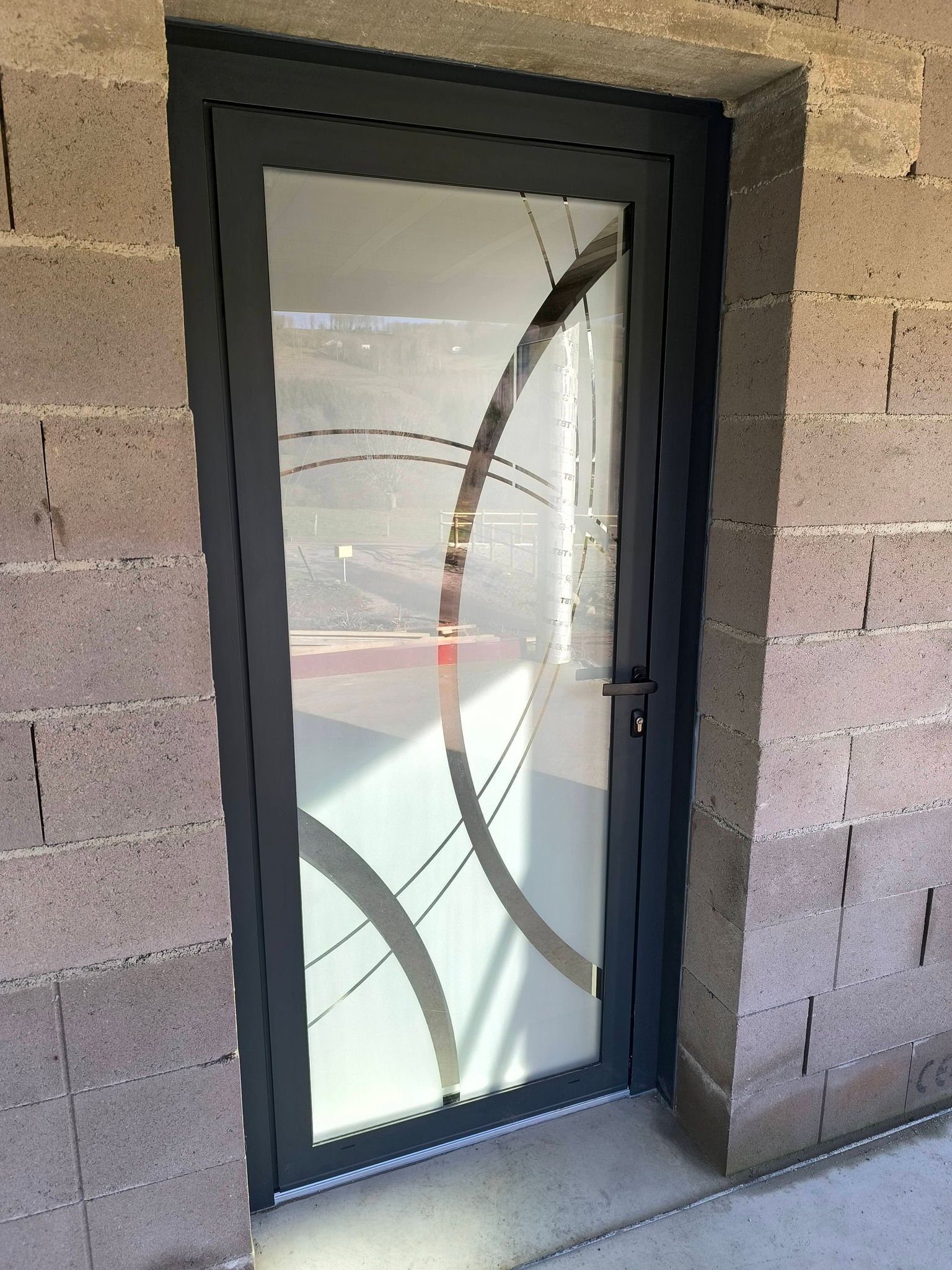 Une porte à cadre gris en aluminium avec un panneau de verre orné d'un motif géométrique circulaire, encastrée dans un mur de parpaings inachevé.