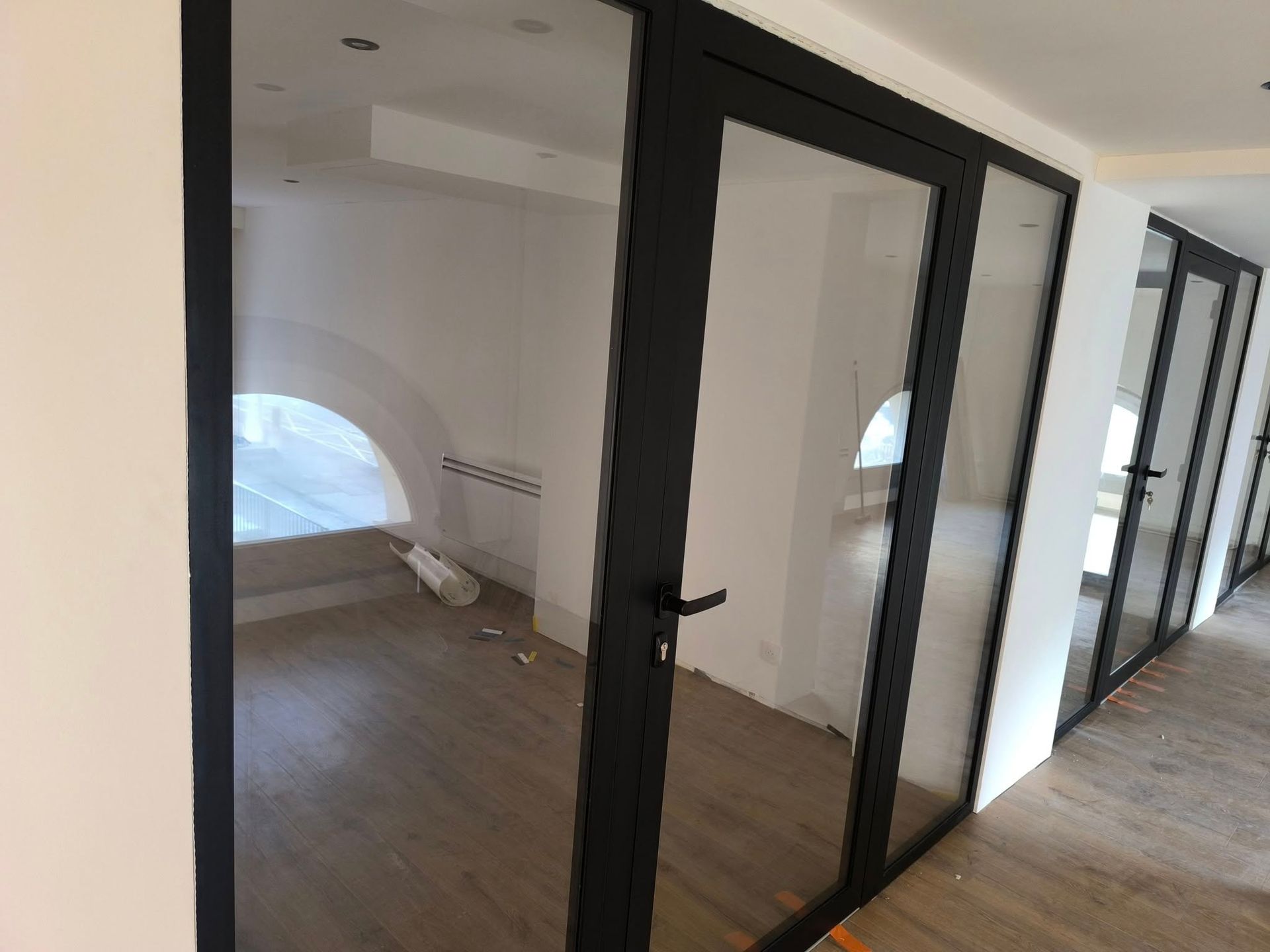 Vue intérieure d'un couloir présentant des portes vitrées à cadre noir menant à des pièces avec des fenêtres cintrées et des parquets.