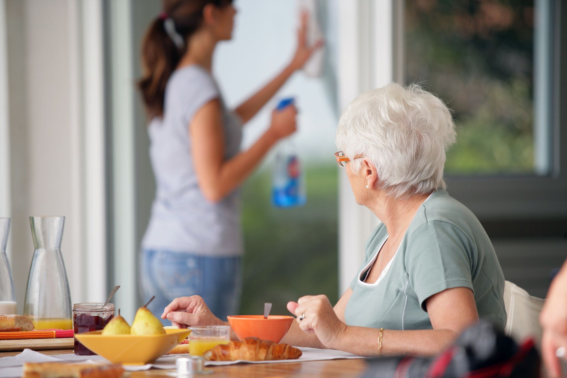 Personne âgée prenant son petit-déjeuner pendant qu'une aide à domicile nettoie les vitres de la baie vitrée