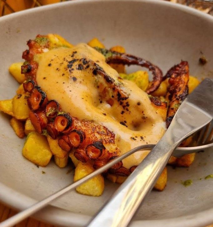 Pulpo a la plancha con patatas, cubierto con salsa, servido en un bol con tenedor y pinzas para servir.