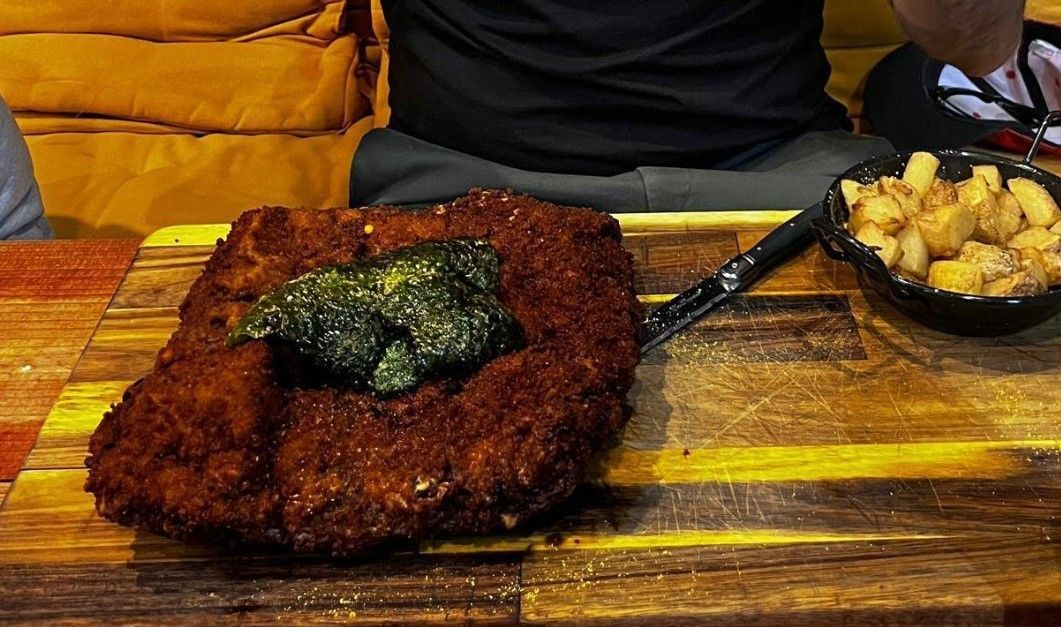 Comida frita grande sobre una tabla de madera con un acompañamiento de comida frita pequeña. Una persona sentada a la mesa.