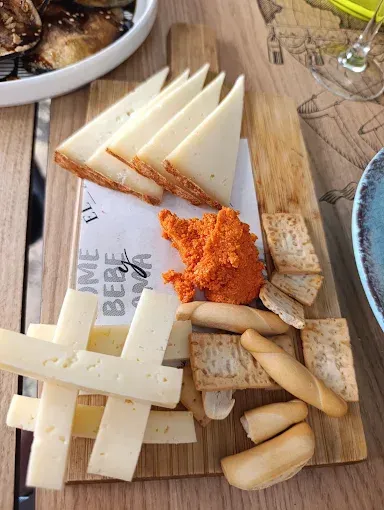 Tabla de quesos con galletas, palitos de pan y pimiento rojo untados sobre una tabla de madera.