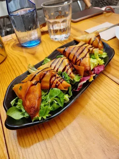 Plato de aperitivos triangulares fritos, bañados en salsa, sobre una base de verduras. Bebidas en una mesa de madera.
