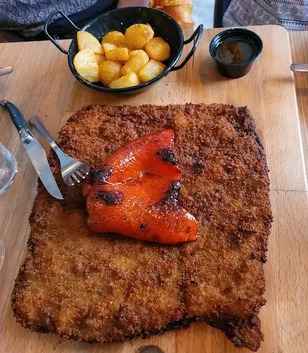 Gran escalope empanado con pimiento rojo asado, patatas y salsa sobre una tabla de madera.