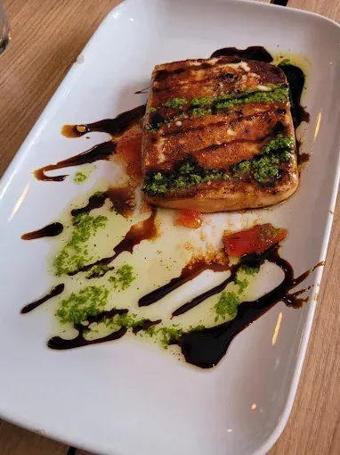 Queso a la parrilla en un plato blanco, rociado con pesto, glaseado balsámico y aceite de oliva.