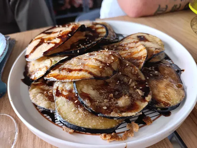 Rodajas de berenjena fritas rociadas con salsa oscura y espolvoreadas con semillas de sésamo en un plato blanco.