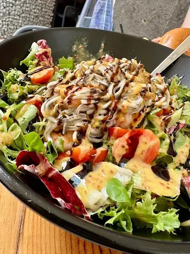 Ensalada en recipiente negro con verduras, tomates, aderezo y una cobertura de fideos blancos.