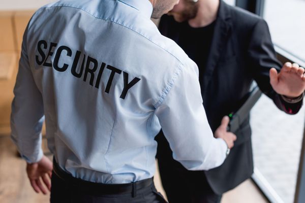 Un guardia de seguridad cachea la chaqueta de un hombre cerca de una puerta. El guardia lleva una camisa azul con la inscripción 