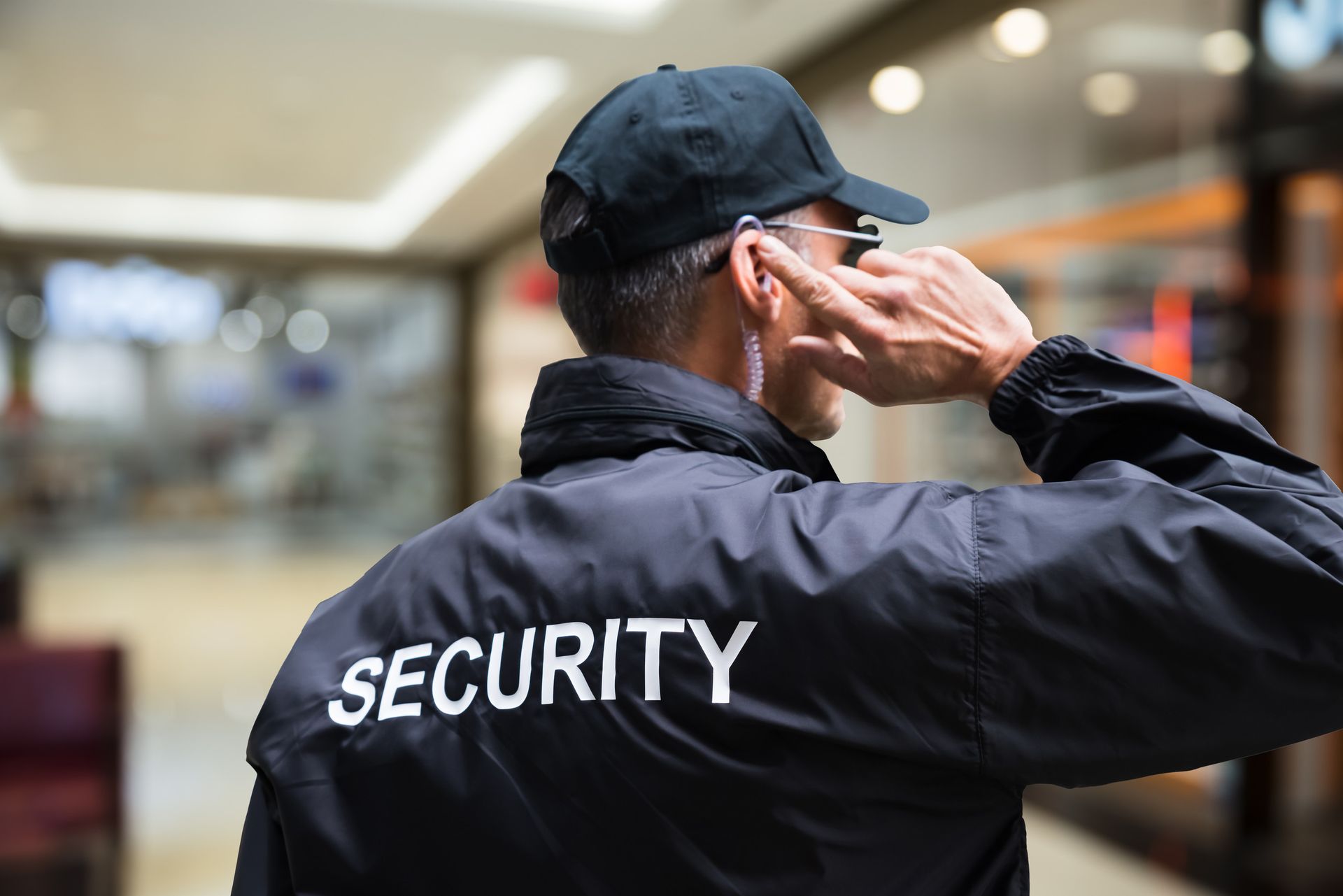 Guardia de seguridad con chaqueta y gorra negras y auricular patrullando el interior de un edificio.