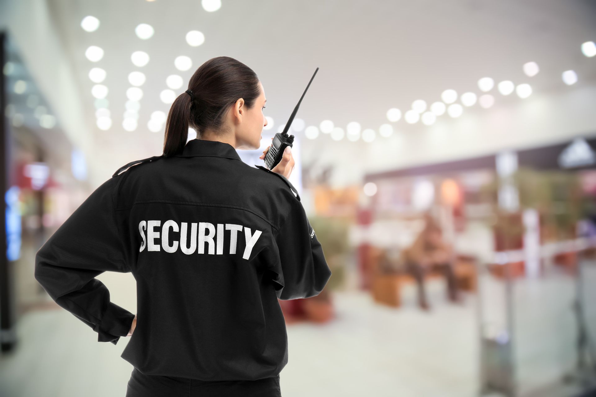 Guardia de seguridad femenina con uniforme negro, sosteniendo una radio, mirando hacia el interior borroso de un centro comercial.