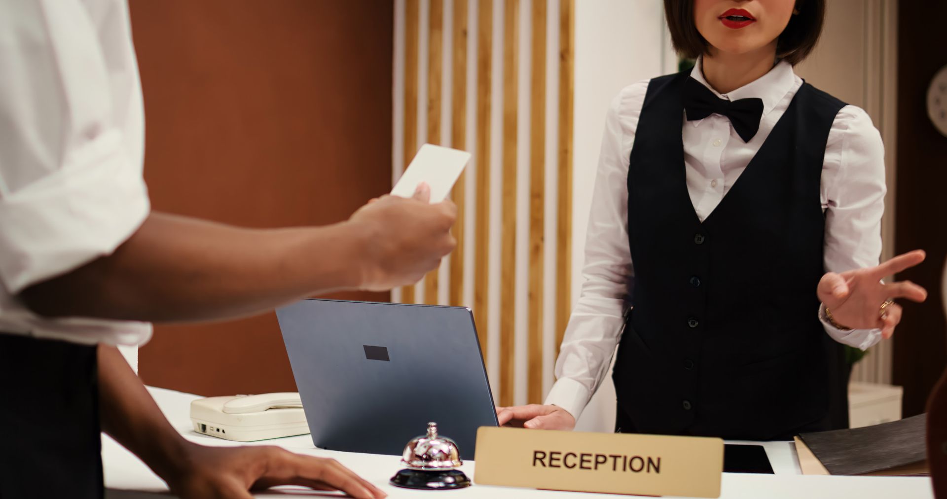 Recepcionista del hotel y huésped en la recepción, huésped sosteniendo una tarjeta, recepcionista haciendo un gesto, computadora portátil y timbre presentes.