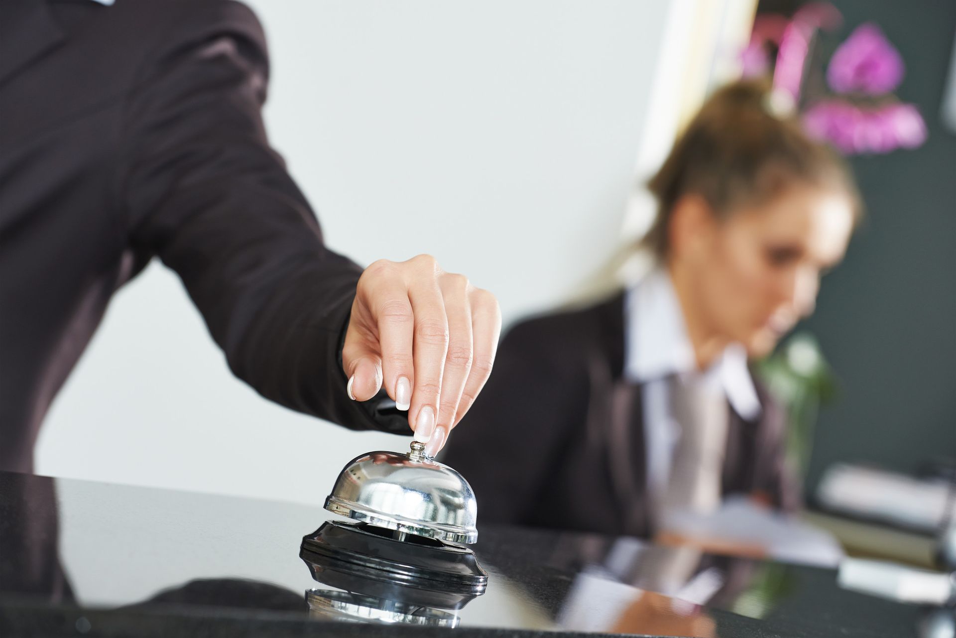 La mano de una persona hace sonar una campana de plata en el mostrador de recepción de un hotel, con un empleado del hotel al fondo.