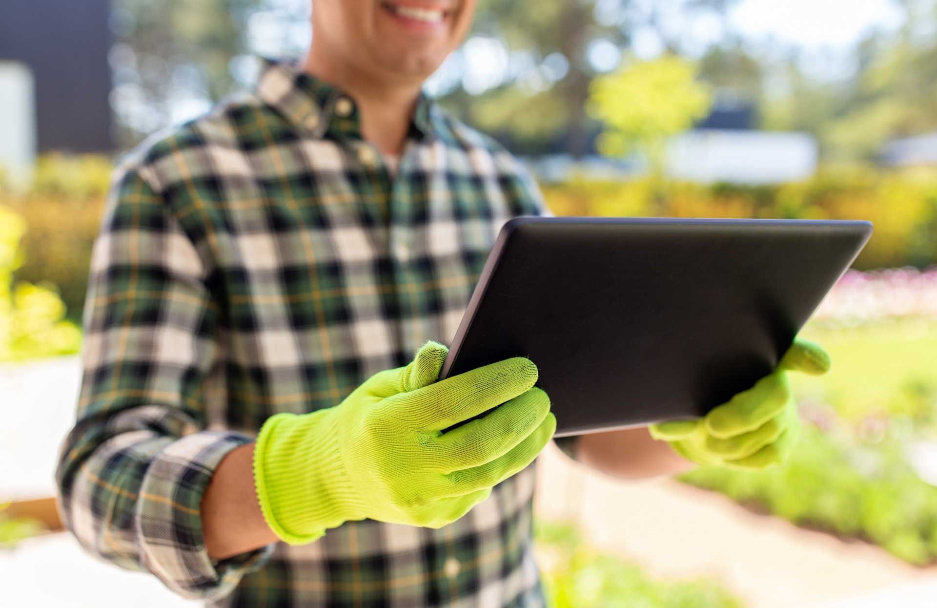Hombre con camisa a cuadros verde y guantes sosteniendo una tableta, sonriendo al aire libre en un jardín.